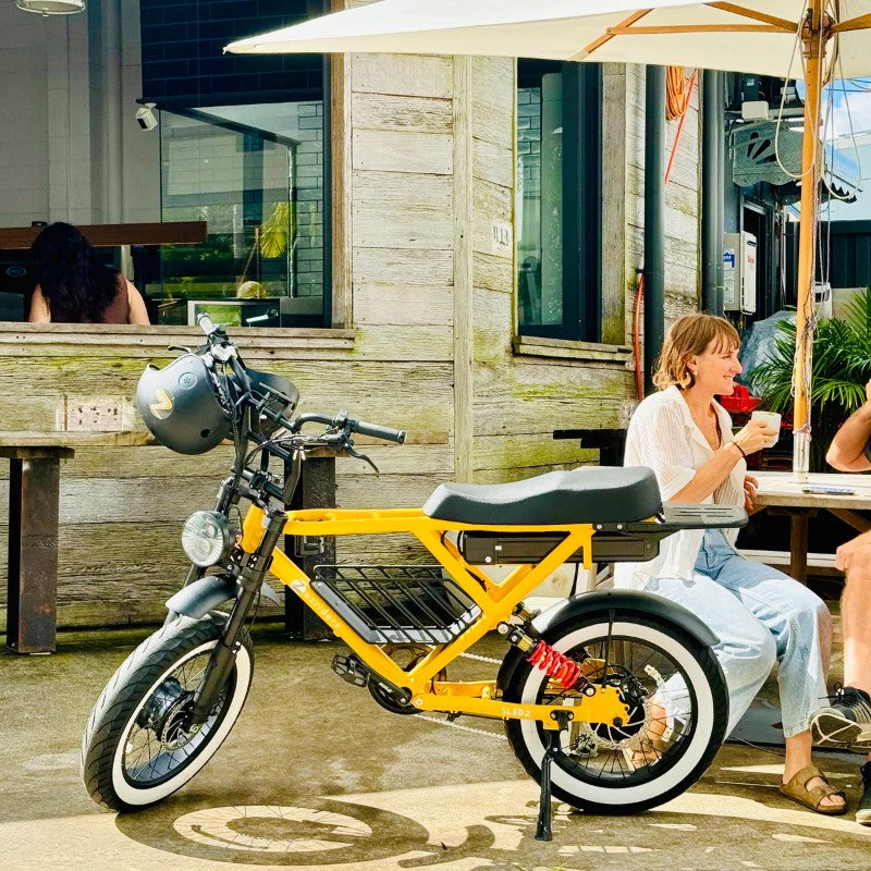 Beast Electric Bike in Colour Spicy Mustard Parked Outside a Rustic Cafe with Two People Casually Enjoying Drinks at a Shaded Outdoor Table